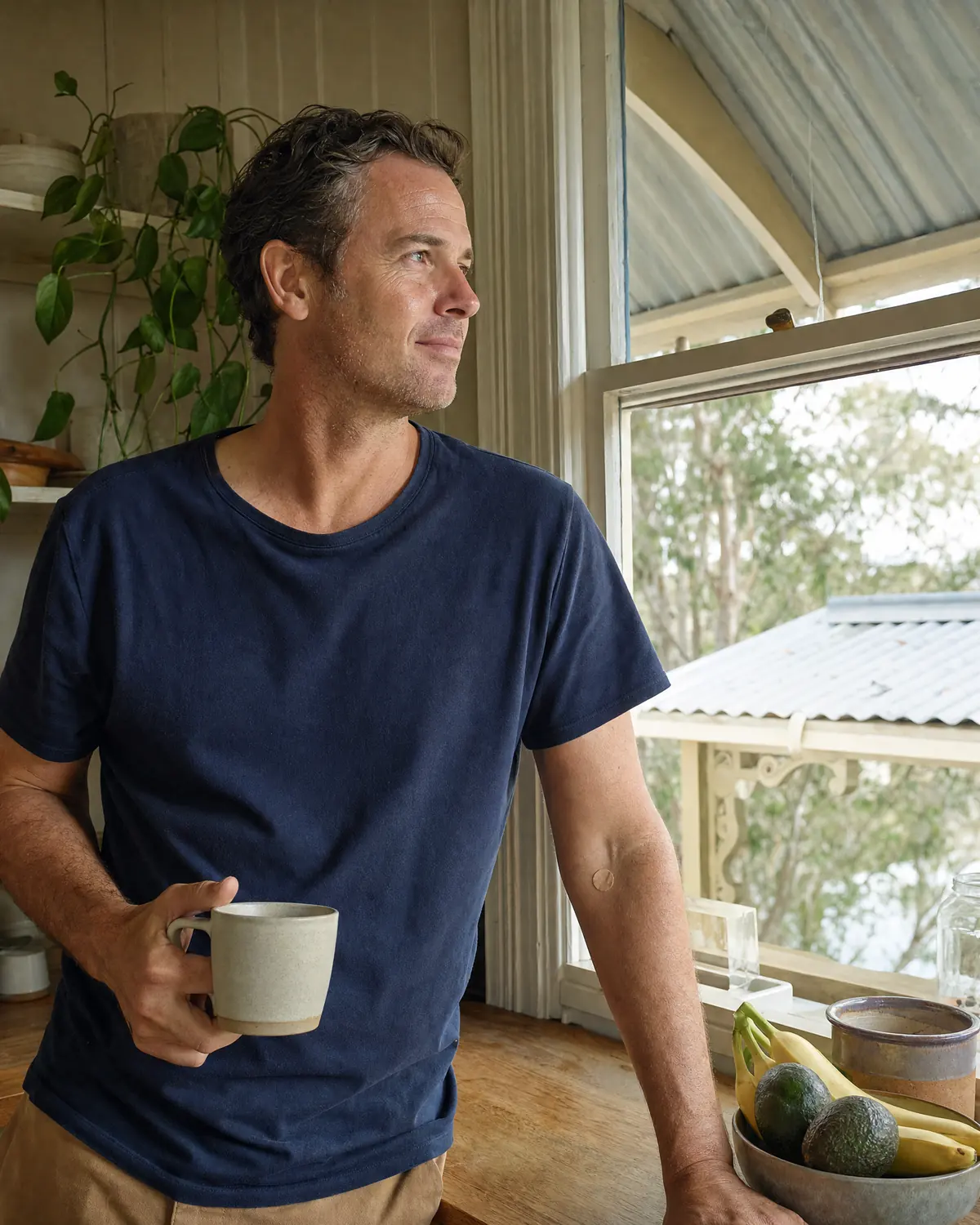 A man in a navy t-shirt holds a mug in a Queenslander kitchen, a small plaster on his inner elbow after a routine blood collection.